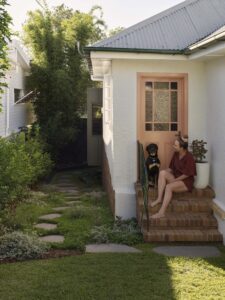 A woman sitting on the home's front brick steps with her dog, showcasing the extension at the rear.